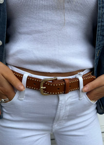 A person wearing a white ribbed shirt tucked into white jeans with a studded brown leather Frame Belt from Frame. Their hands are tucked into the jeans' pockets, and a denim jacket can be seen on their sides. Gold rings are visible on their fingers.