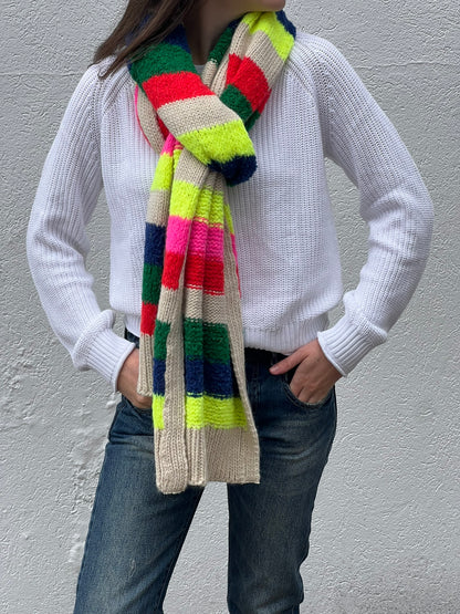 A person wearing a white knitted sweater and blue jeans is wrapped in an Essentiel Antwerp Garis Knitted Scarf, featuring vibrant shades of green, yellow, pink, red, and black. The heavyweight knit scarf showcases bold intarsia-knit patterns as they stand against a plain textured wall with their hands in their pockets.