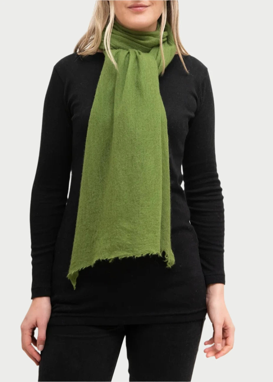 A person with shoulder-length blonde hair is standing against a white background, wearing a luxurious bright green Whisperweight Cashmere Scarf by Meg Cohen around their neck over a black long-sleeve top and black pants.