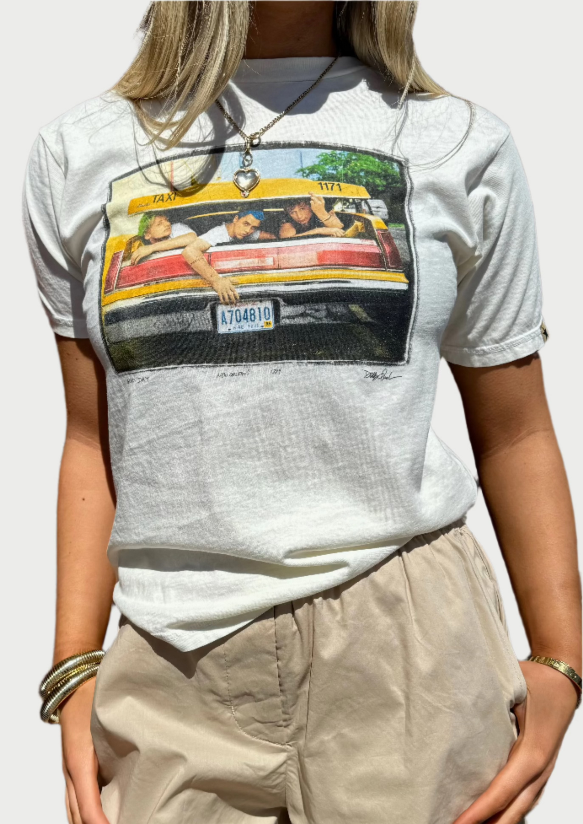 A person wearing a CLINCH Concert Tees depicting a group of people in the back of a taxi and beige shorts stands against a brick wall. The combed cotton outfit is accessorized with gold bracelets and a necklace. The person has long, blonde hair.