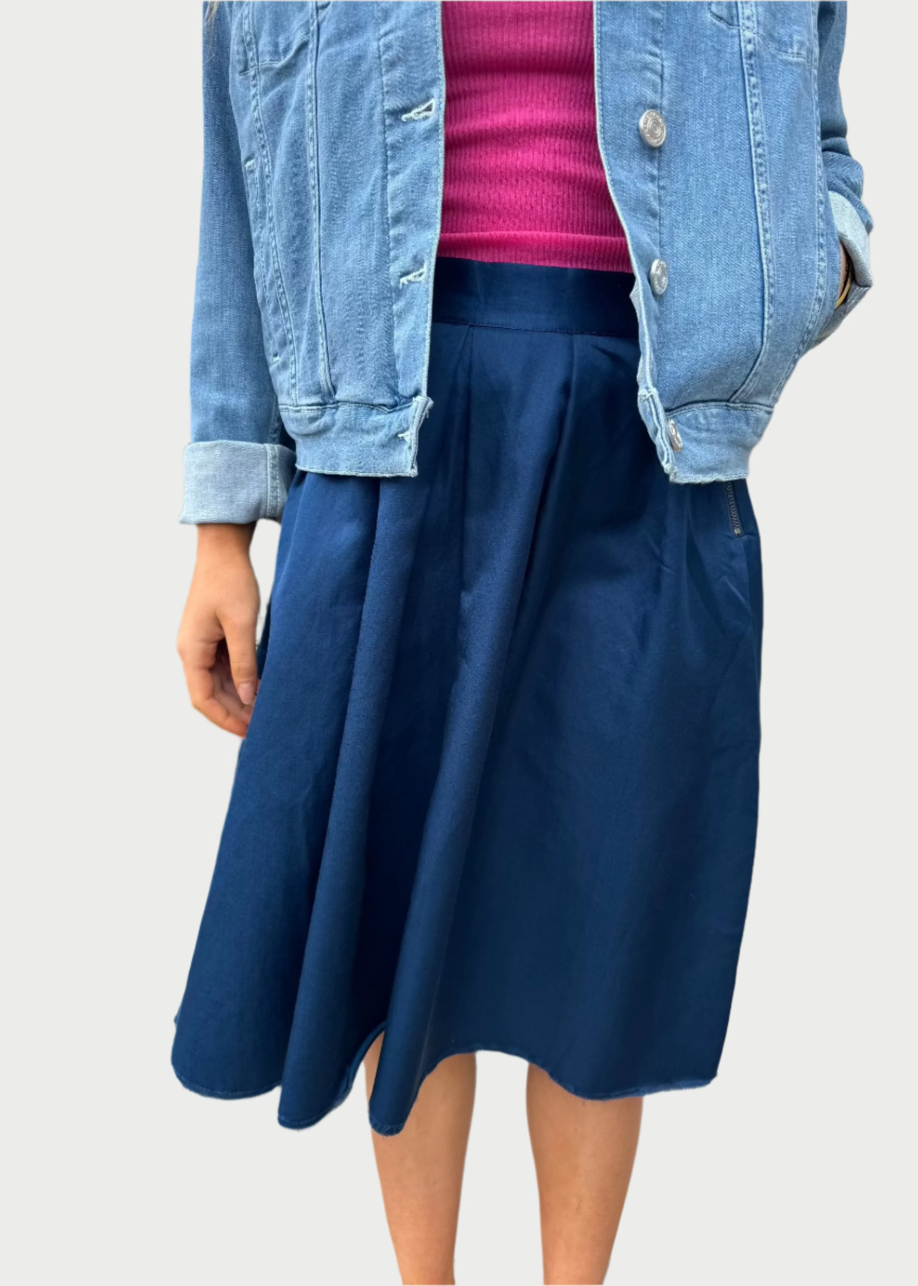 A person stands outdoors in front of greenery and a white wall, wearing a light blue denim jacket over a hot pink top, paired with a dark blue high-waisted Double Pleated Skirt from Brazeau Tricot. The image focuses on their torso and waist, showcasing a flattering silhouette with their face not visible.