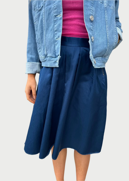 A person stands outdoors in front of greenery and a white wall, wearing a light blue denim jacket over a hot pink top, paired with a dark blue high-waisted Double Pleated Skirt from Brazeau Tricot. The image focuses on their torso and waist, showcasing a flattering silhouette with their face not visible.