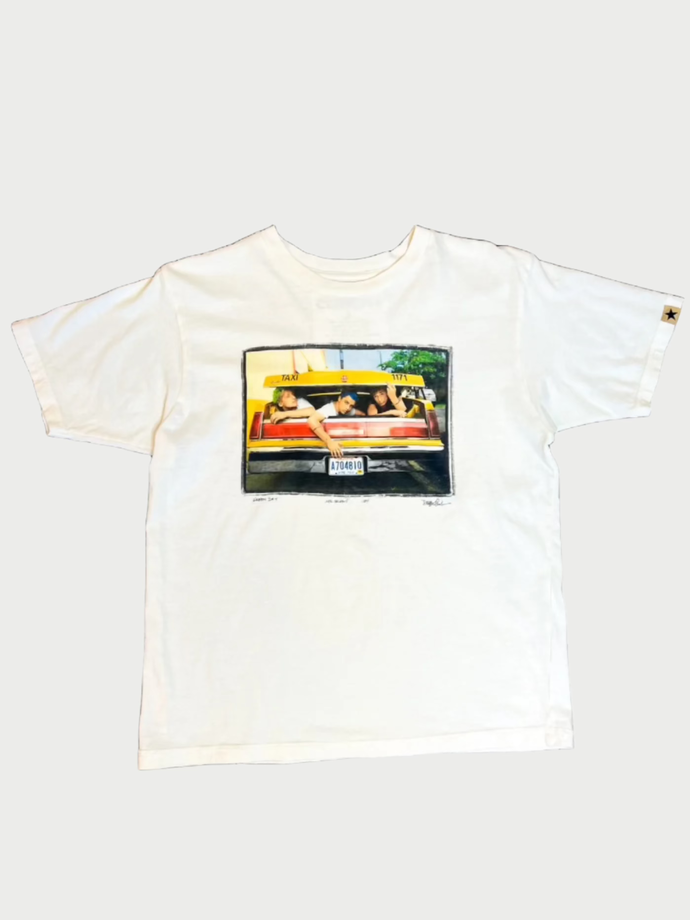 A vintage-washed white **Concert Tees** featuring a colorful graphic by photographer Danny Clinch of a group of people lounging in a car, with trees in the background. The license plate of the car is visible at the bottom center of the image. Made from premium combed cotton, this short-sleeved tee has a crew neck design by **CLINCH**.