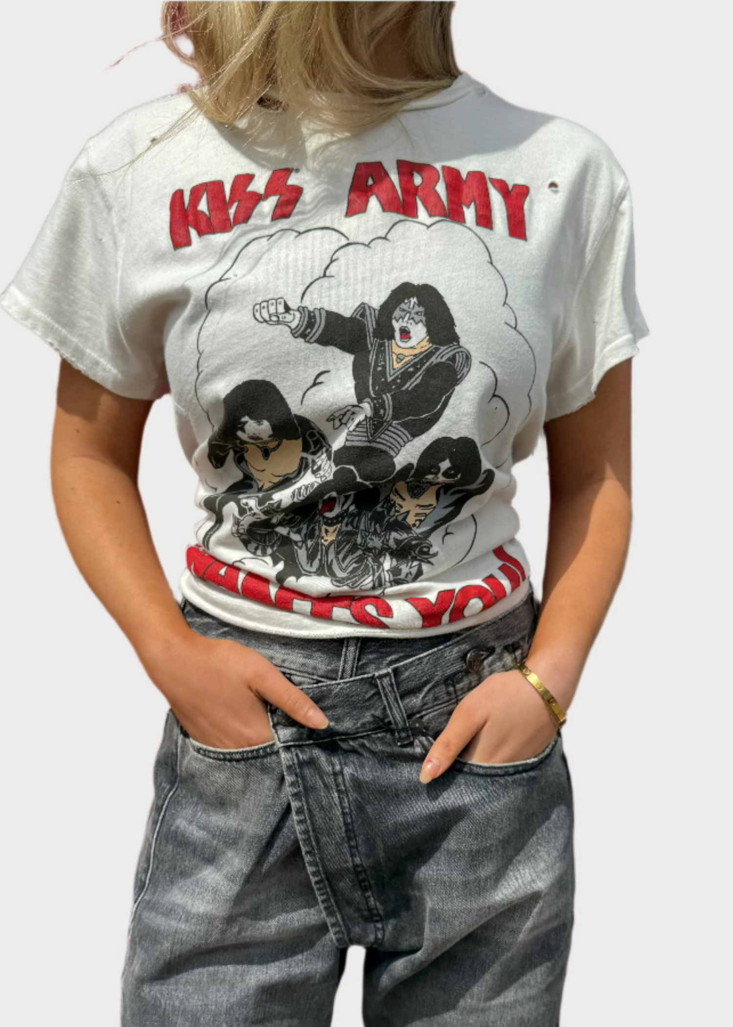 A person is standing against a bright red door, wearing a white "KISS Destroyed Tee" by Madeworn featuring cartoon characters of the band members, and gray high-waisted jeans with a relaxed fit. Their hands are in their pockets, and their face is not visible in the frame.