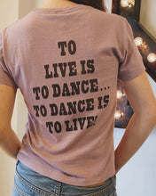 Load image into Gallery viewer, A person wearing a vintage-inspired Snoopy Dancing Classic Tee from RE/DONE, featuring a relaxed fit and the message &quot;TO LIVE IS TO DANCE... TO DANCE IS TO LIVE!&quot; printed in black on the back. The light purple shirt creates a stylish look as the person stands indoors near a lit-up marquee letter &quot;A.