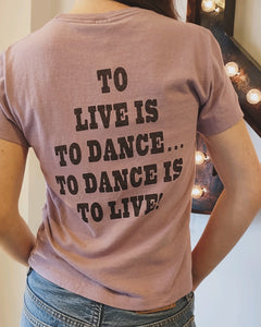 A person wearing a vintage-inspired Snoopy Dancing Classic Tee from RE/DONE, featuring a relaxed fit and the message "TO LIVE IS TO DANCE... TO DANCE IS TO LIVE!" printed in black on the back. The light purple shirt creates a stylish look as the person stands indoors near a lit-up marquee letter "A.