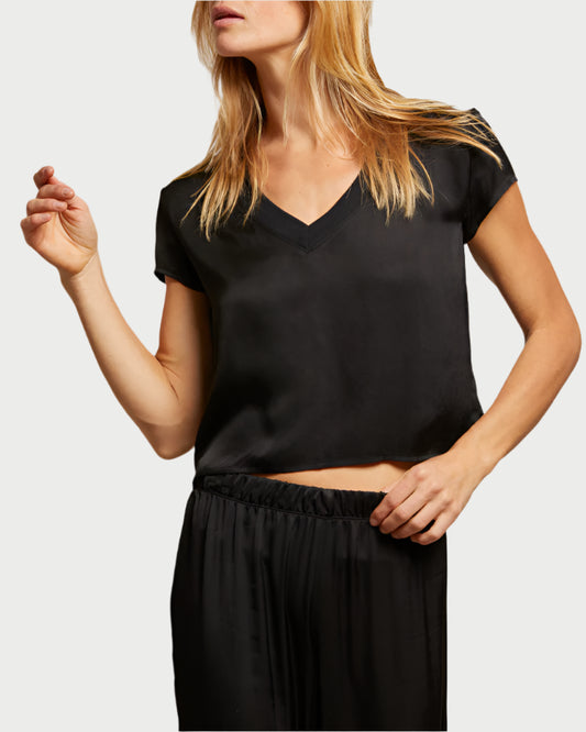 A person wearing the perfectwhitetee Amelie Satin V Neck Tee in black with matching pants stands against a light background. Their blonde hair and slight side turn highlight the sleek ensemble, crafted from 100% viscose.