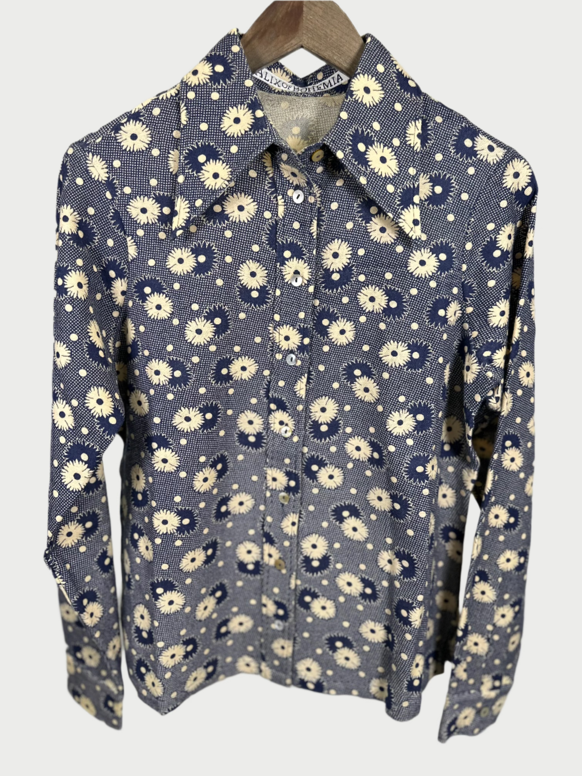 The Daisy Print Shirt by Alix of Bohemia features long sleeves and a vibrant blue background adorned with small white and yellow floral patterns, evoking the essence of fall. Displayed elegantly on a wooden hanger against a simple backdrop, this button-down shirt captivates with its stylish design.