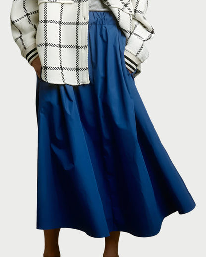 A person in a white and black plaid jacket with hands in its pockets pairs it with AqC's Le Skirt, a long taffeta skirt featuring an elastic waist and a slight sheen, standing against a plain background.