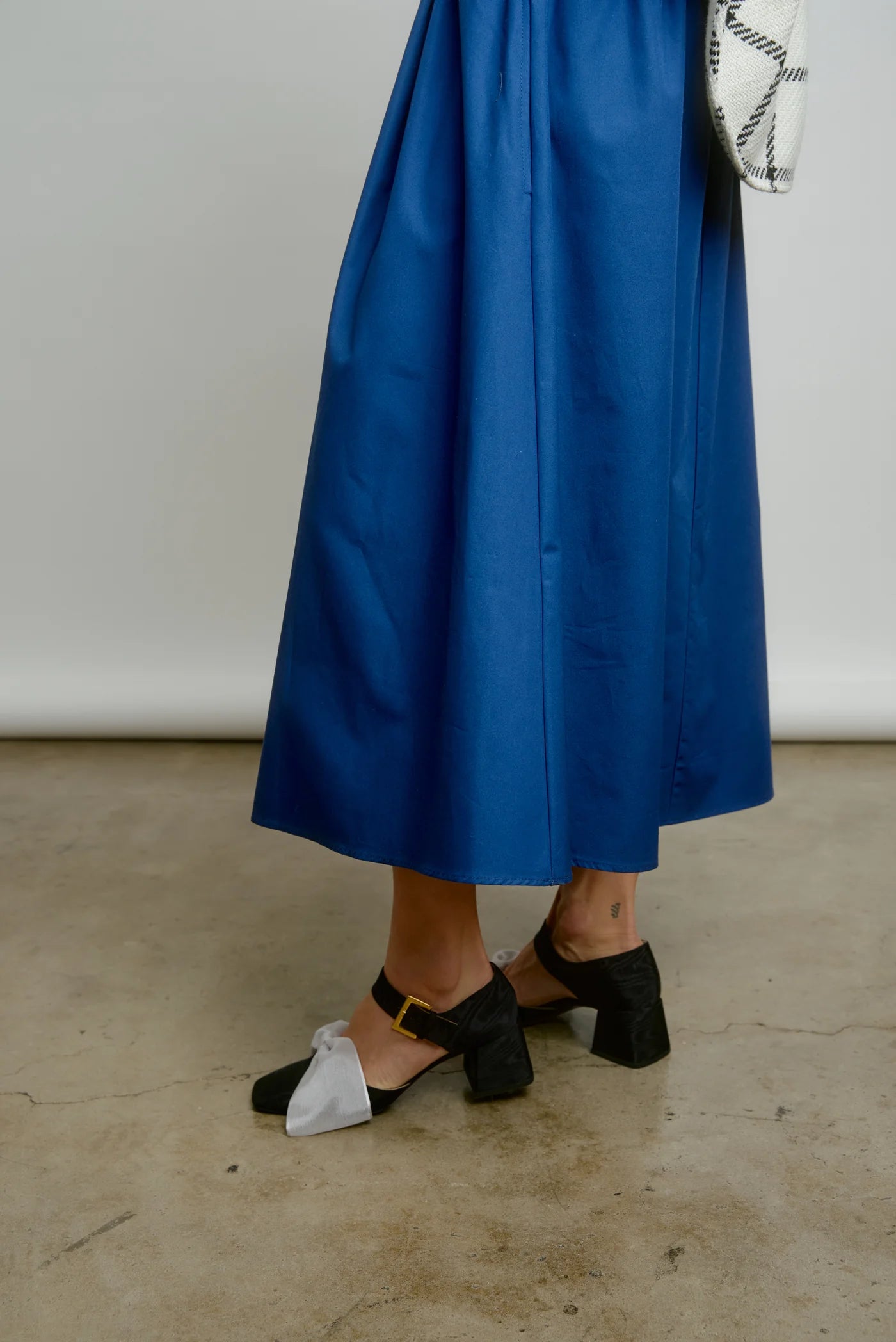 A person wearing AqC's Le Skirt, a long blue taffeta skirt with an elastic waist, and black heeled shoes with white toe caps stands on a concrete floor while holding a patterned handbag against a plain, light-colored wall.