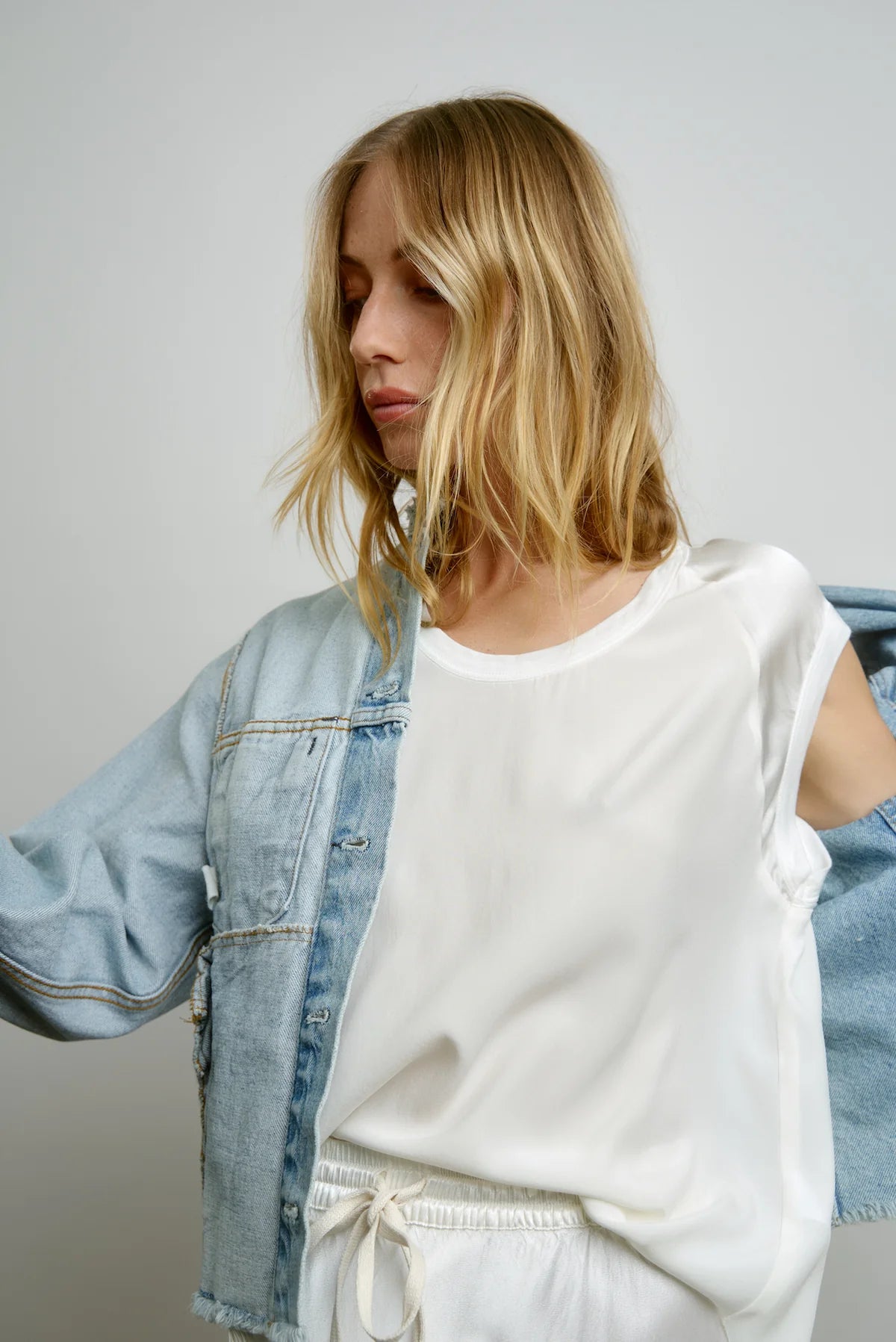 A woman with blonde hair is wearing the Cap Sleeve Silk Baseball Tee in Cloud by AqC, paired with white pants and partially covered by a light wash denim jacket featuring shiny sleeves. She stands against a plain background, looking downward with her arms slightly raised.