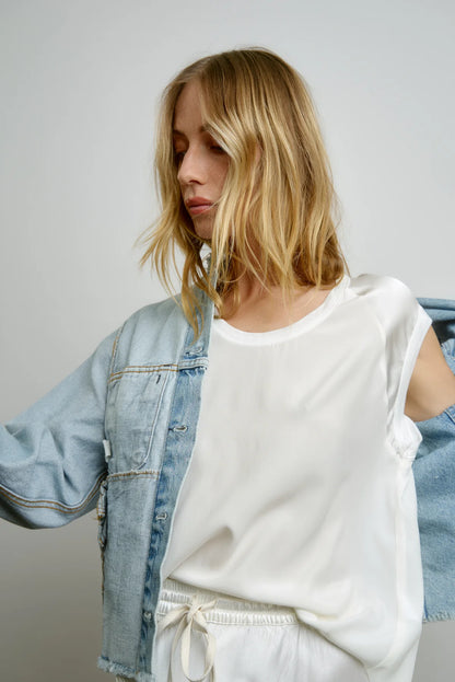 A woman with blonde hair is wearing the Cap Sleeve Silk Baseball Tee in Cloud by AqC, paired with white pants and partially covered by a light wash denim jacket featuring shiny sleeves. She stands against a plain background, looking downward with her arms slightly raised.