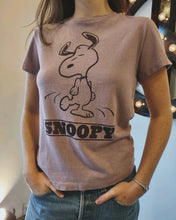 Load image into Gallery viewer, A person wearing the Snoopy Dancing Classic Tee by RE/DONE, a vintage-inspired purple t-shirt featuring an illustration of Snoopy from the Peanuts comic strip dancing with &quot;Snoopy&quot; written below. The relaxed fit of the shirt ensures they stand confidently with one hand in their pocket, while part of a decorative light is visible in the background.