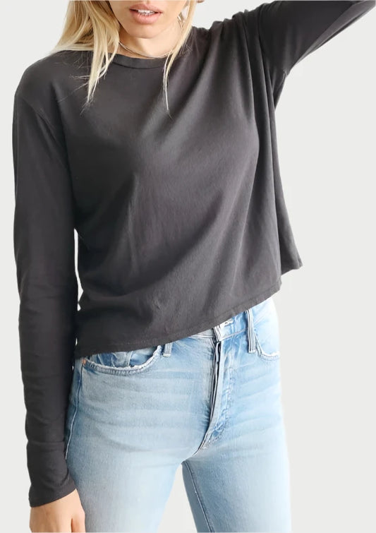 A person with long hair wears a Long Sleeve Tee black top and light blue jeans. They have one hand behind their head and are looking slightly downward. The background is a plain white wall, making their perfectwhitetee stand out effortlessly.