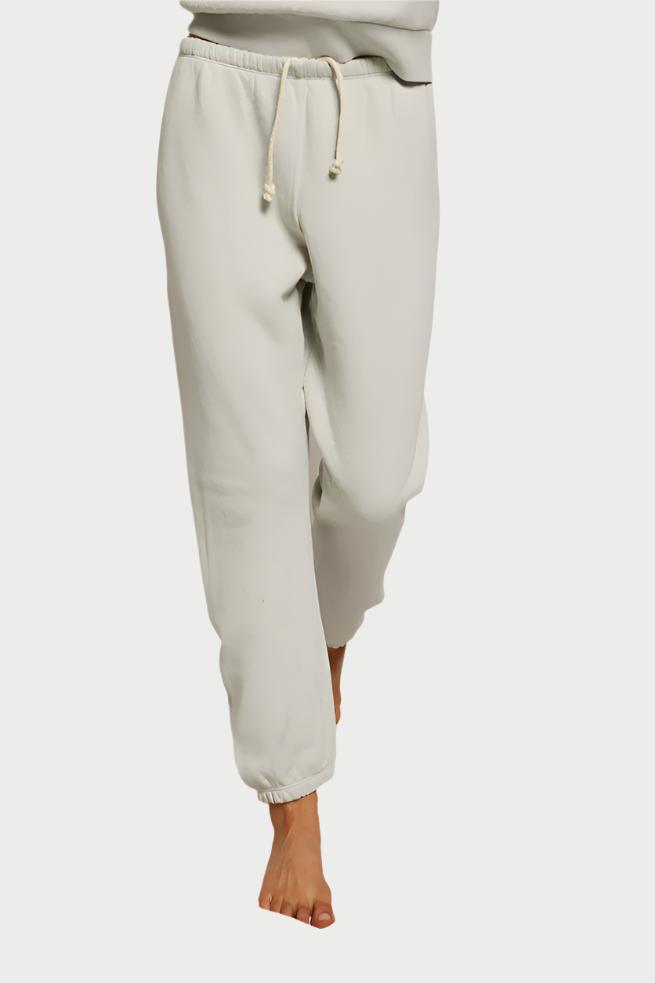 A person wearing light-colored, loose-fitting perfectwhitetee Stevie Sweatpant joggers with a hidden drawcord stands barefoot on a soft, crumpled white background. The person's upper body is partially out of frame, and the joggers have gathered cuffs at the ankles.