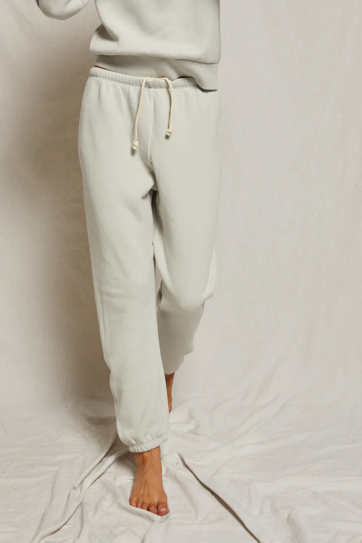A person wearing light-colored, loose-fitting perfectwhitetee Stevie Sweatpant joggers with a hidden drawcord stands barefoot on a soft, crumpled white background. The person's upper body is partially out of frame, and the joggers have gathered cuffs at the ankles.