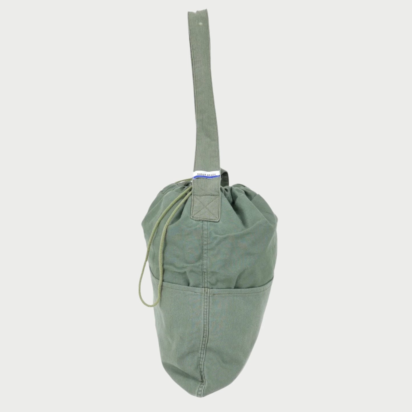 A green SUPER READY Drawstring Tote, crafted from up-cycled Vintage US Military fabric, hangs by its loop handle. The bag boasts a spacious lower compartment and a cinched top, along with an adjustable shoulder strap for versatile carrying options.