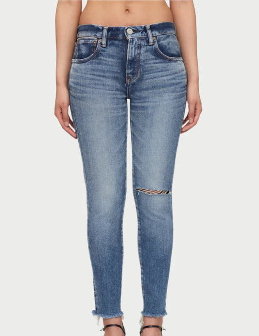 A person is wearing Moussy Lawrenceville Jeans, mid-rise, light blue skinny jeans with a distressed patch on one knee and frayed hems. The vintage-inspired jeans are paired with black, open-toed heels. The image is cropped at the waist and feet, showing only the lower half of the body against a plain background.