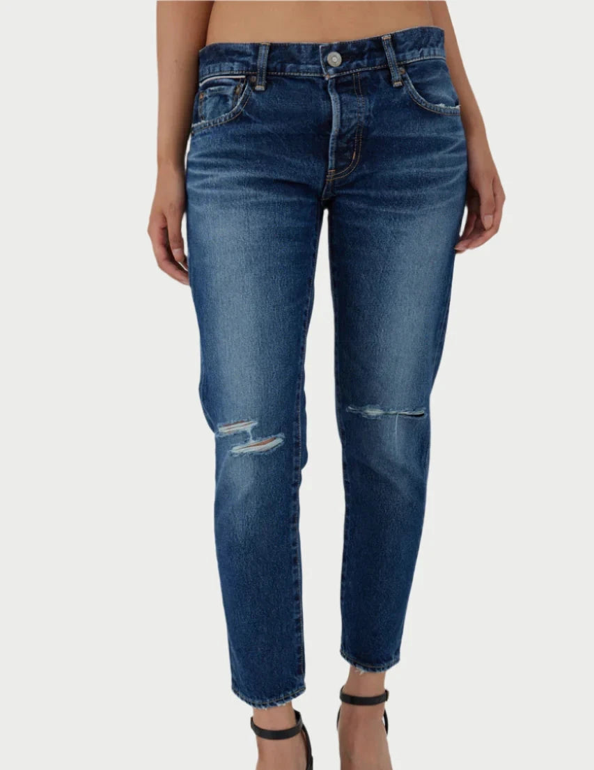 A person is modeling a pair of Moussy Rosemead Tapered Jeans in blue, featuring rips on the left knee and slight fraying on the right thigh. These mid-rise jeans come with a button and zipper closure, along with front and back pockets. They are paired with black strappy sandals against a plain white background.