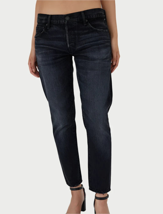 A person wearing Moussy's Vellflower Jeans, featuring a high-rise waist and relaxed fit in black denim, paired with black ankle-strap heels. The background is a plain light gray.