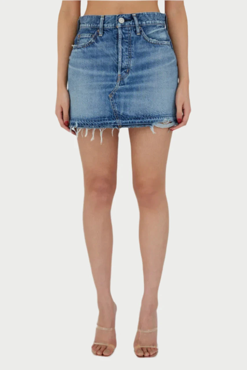 A person is wearing the Mini Denim Skirt by Moussy, which is high-waisted with frayed edges, visible front pockets, and a zipper. The skirt boasts a raw hemline and is paired with clear heeled sandals, making it an ideal choice for a casual lunch outfit. The background is plain white.