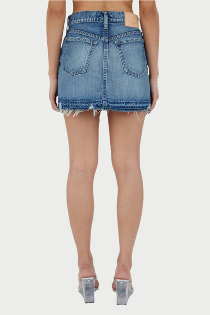 A person is standing with their back facing the camera, showcasing a frayed blue Mini Denim Skirt by Moussy and clear high-heeled shoes. The plain white background accentuates the casual outfit, highlighting the skirt's two back pockets and raw hem.