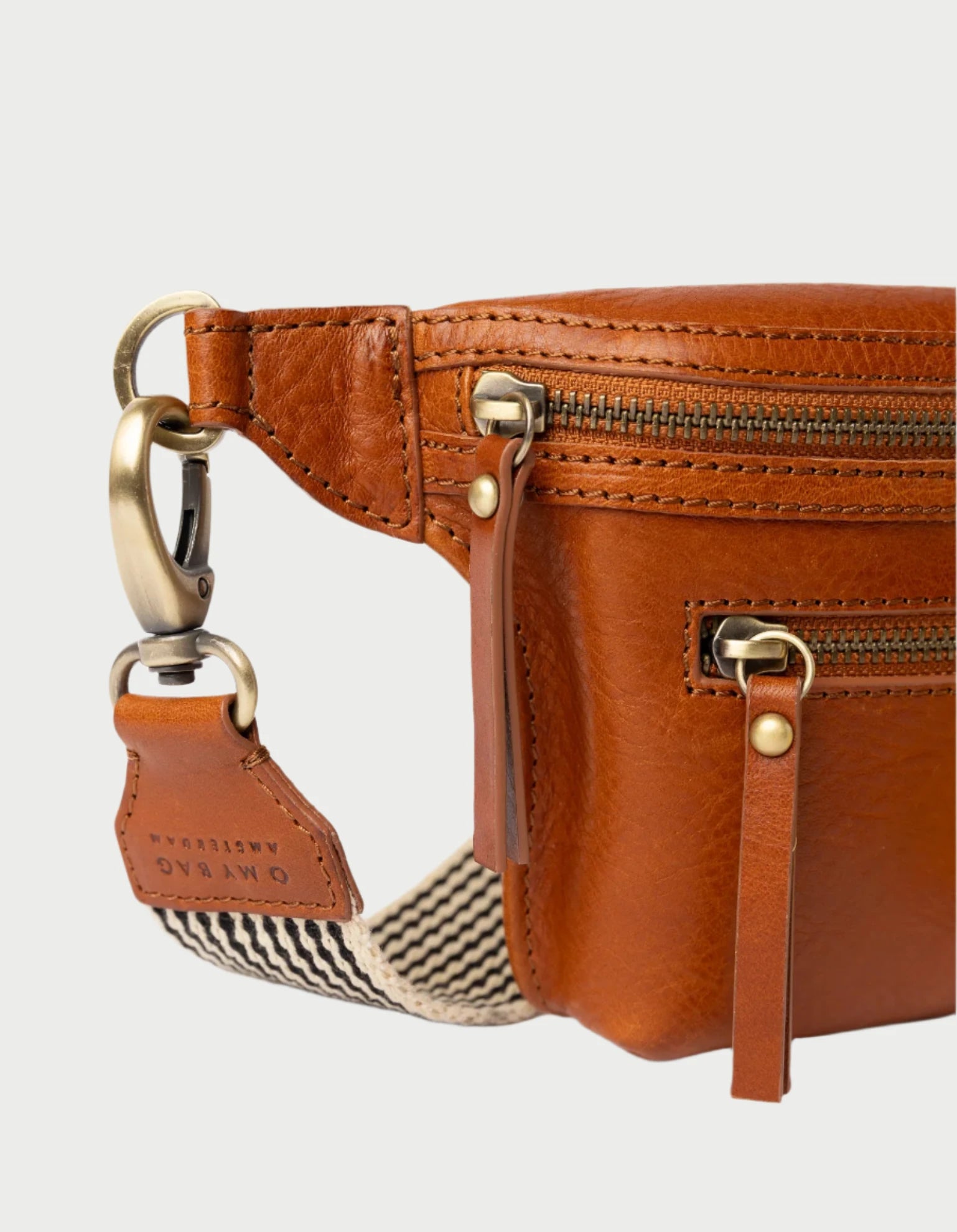 Close-up of Beck's Bum Bag in Cognac by O MY BAG. The bag is crafted from brown leather and is accented with a brass zipper and clasp. It features a woven strap with stripes, complemented by a leather accent embossed with "OYABAO." Its vintage-inspired design includes visible stitching and leather pulls.