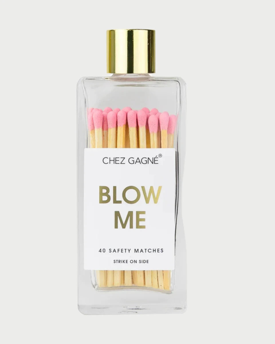 A glass bottle holds 40 colorful wooden matches, each featuring a pink tip and finished with a gleaming gold cap. The label displays "Chez Gagné Blow Me," accompanied by the text "40 Safety Matches Strike On Side.