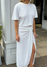 Load image into Gallery viewer, A person stands outdoors wearing a white, short-sleeved, **Le Superbe White Party Sequin Tee** paired with a high-waisted, ankle-length skirt featuring a side slit. The background shows a building with grey paneling and a glass door. The person&#39;s head is not visible in the image.
