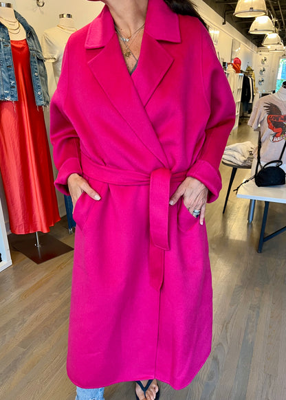 A person wearing a vibrant fuchsia Double Face Coat by Essentiel Antwerp stands in a clothing store. The bright pink, belted coat features a wide collar and large front pockets. In the background, various clothing items like a red dress and denim jacket are visible.