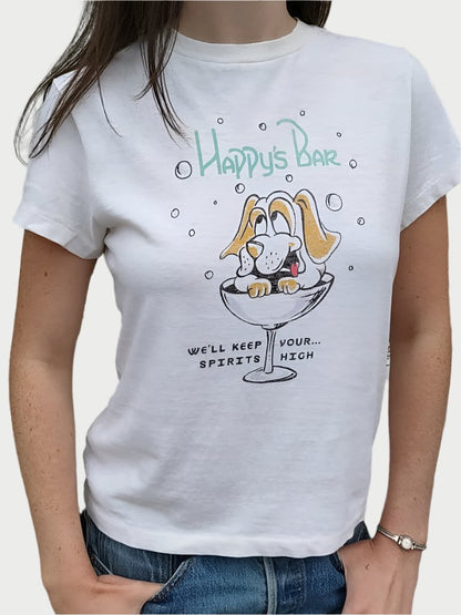 A person wears the RE/DONE Happys Classic Tee, made of ultra-soft cotton, featuring a cartoon dog in a cocktail glass, smiling with paws on the rim. Text above the dog reads "Happy's Bar," while below it says, "We'll keep spirits high." The vintage-inspired design and quality fabric make it a standout piece. Plants and flowers are visible in the background.