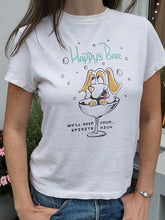 Load image into Gallery viewer, A person wears the RE/DONE Happys Classic Tee, made of ultra-soft cotton, featuring a cartoon dog in a cocktail glass, smiling with paws on the rim. Text above the dog reads &quot;Happy&#39;s Bar,&quot; while below it says, &quot;We&#39;ll keep spirits high.&quot; The vintage-inspired design and quality fabric make it a standout piece. Plants and flowers are visible in the background.