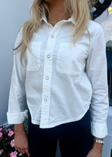 Load image into Gallery viewer, A person is wearing a RE/DONE Slim Denim Shirt in premium cotton with two breast pockets, paired with black pants. Their sleeves are rolled up to the elbows. The person has long, light brown hair and is standing in front of a plain background with some pink flowers visible at the bottom left.