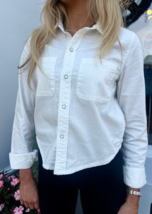 A person is wearing a RE/DONE Slim Denim Shirt in premium cotton with two breast pockets, paired with black pants. Their sleeves are rolled up to the elbows. The person has long, light brown hair and is standing in front of a plain background with some pink flowers visible at the bottom left.