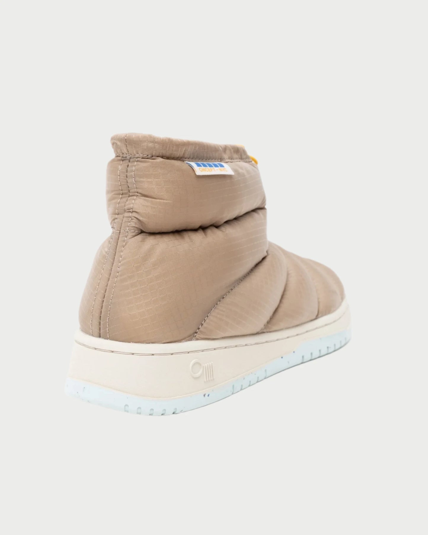 Introducing the Calgary Bootie by Oncept: this beige puffer sneaker boot showcases a quilted texture and a molded footbed. Enhancing comfort with its thick sole, it also features a label on the side. The shoe is presented from the back against a simple light background.