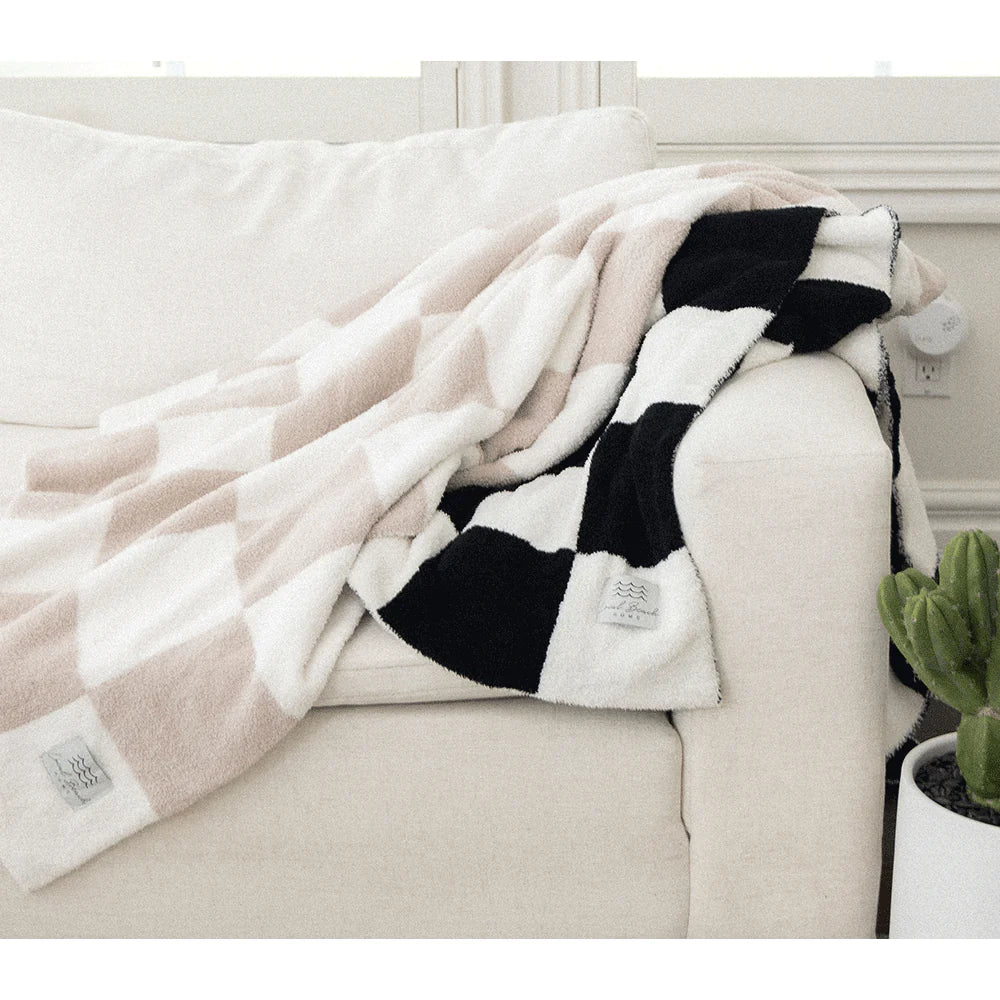 A cozy living room scene features a cream-colored sofa with the XL LOCAL BEACH Luxe Checkered Blanket in black and white draped over the arm. Made from soft polyester microfiber, it complements the small potted cactus on the floor nearby, adding a touch of greenery.