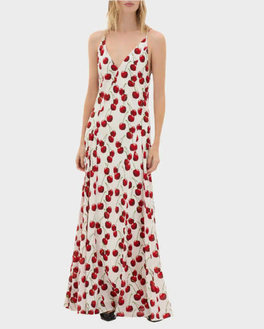 A person is elegantly dressed in the Altair Cherries Dress by MUMA, featuring a V-neckline and a flowing skirt with red cherry patterns. With shoulder-length hair, they pose gracefully against a plain, light background.