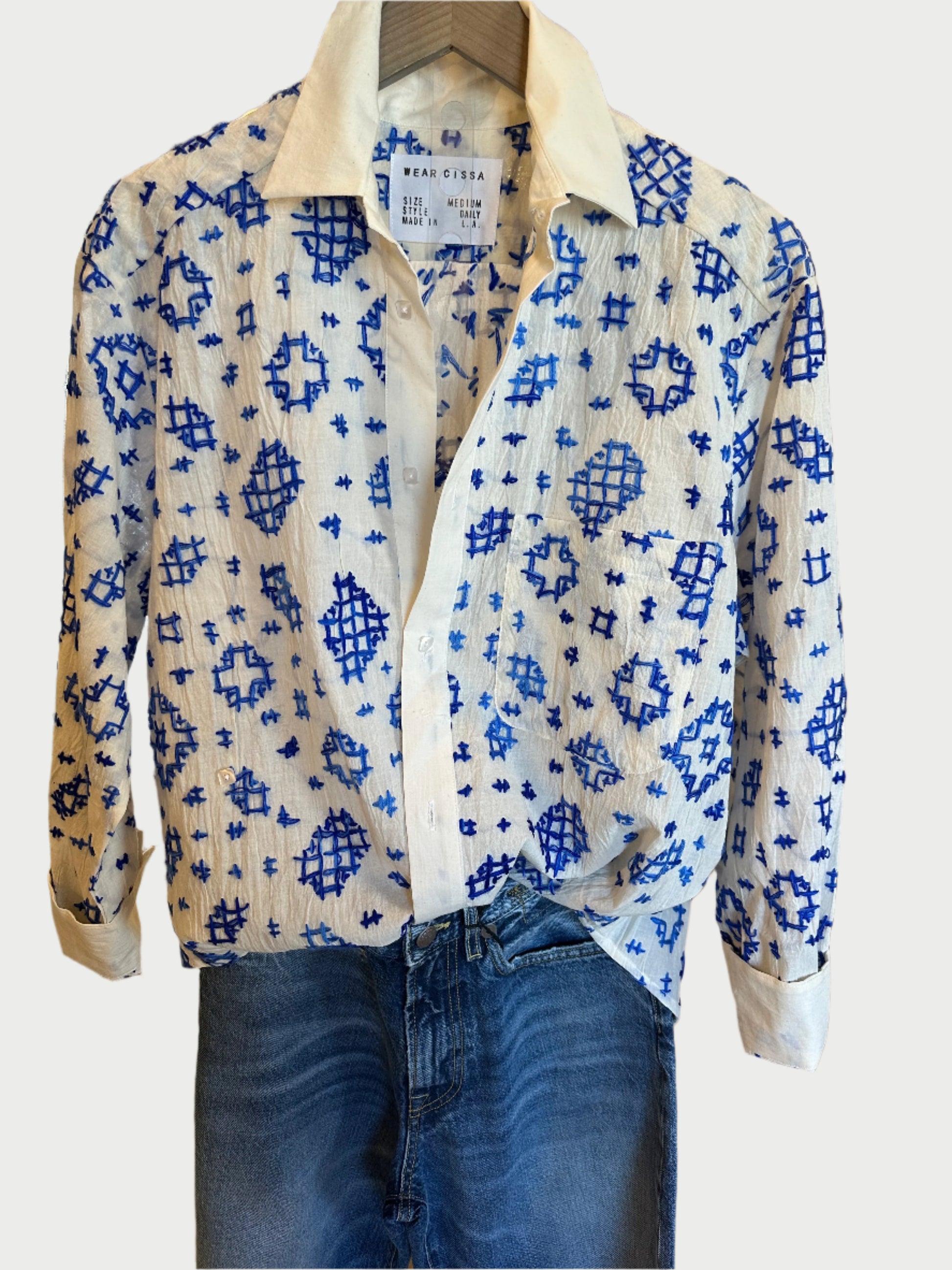 The Daily Cotton Shirt by WEAR CISSA, a white button-up with blue geometric patterns, relaxed fit, collar, and long sleeves, hangs above classic-textured blue jeans. Both garments are elegantly showcased against a plain backdrop.