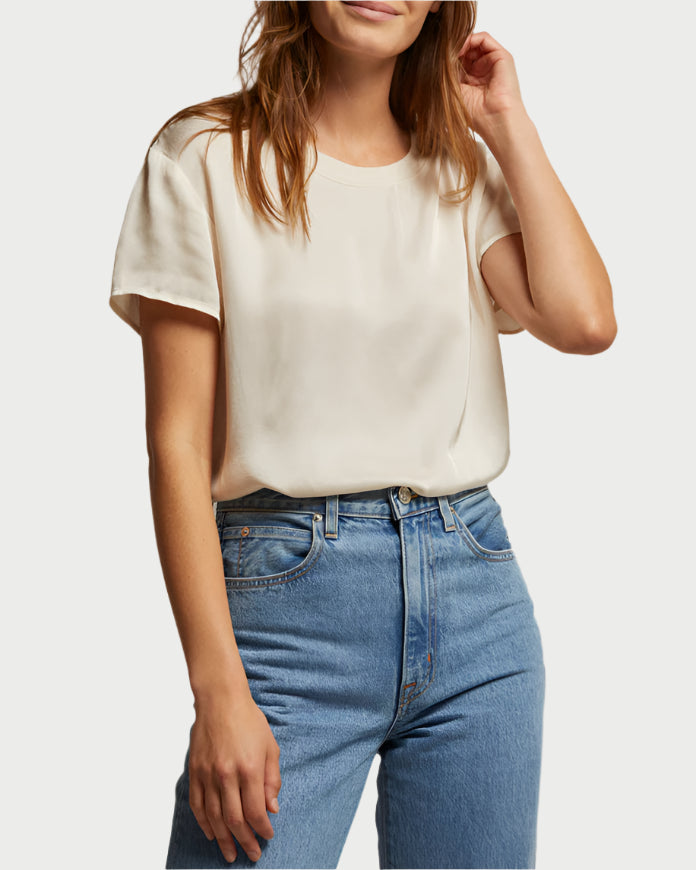 Wearing a perfect white tee Delphine Satin Crewneck in cream and blue high-waisted jeans, the person stands against a plain backdrop. They have long brown hair and raise their left hand to their ear.