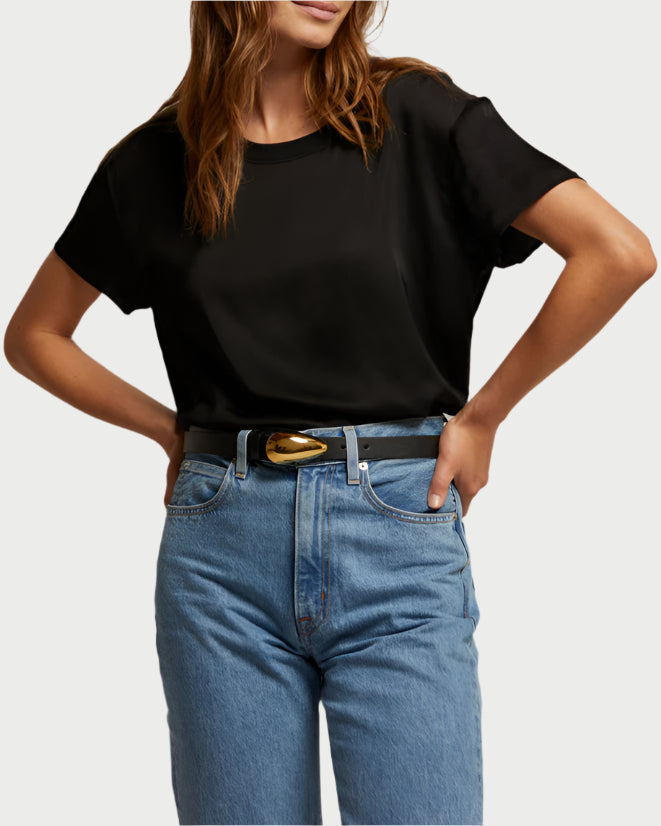 A person wearing a black Delphine Satin Crewneck Tee from perfect white tee and blue jeans, with hands on hips. A stylish belt featuring a gold buckle adds an elevated touch against the plain background.