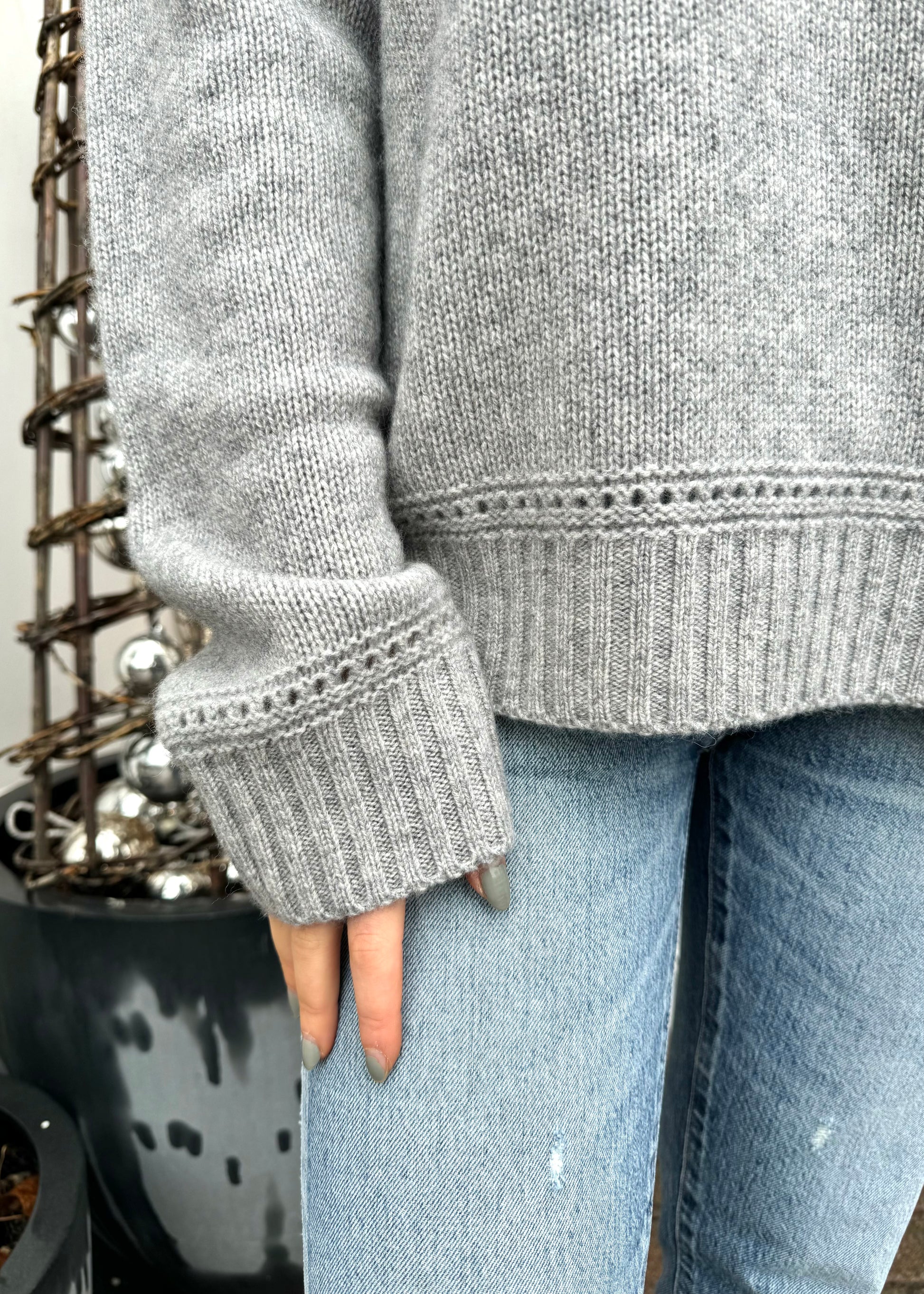 A person is wearing a cozy grey Karyna Cashmere Sweater by One Grey Day with detailed ribbed cuffs and hem, paired with light blue denim jeans. The background features a potted plant with a decorative, rustic arrangement, adding a touch of natural, festive charm.