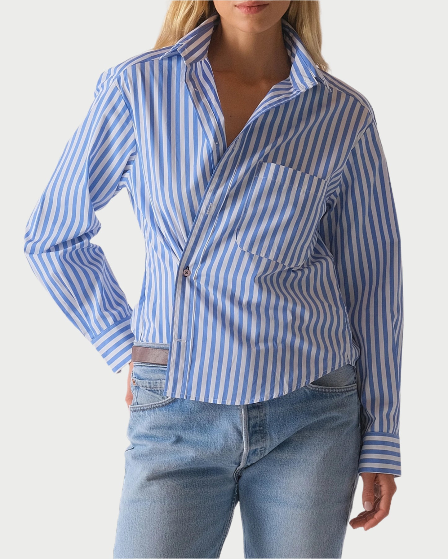 In a plain backdrop, someone wears The Daily Shirt by WEAR CISSA, featuring blue and white vertical stripes in luxurious Egyptian cotton, paired with blue jeans and one hand casually placed in their pocket.