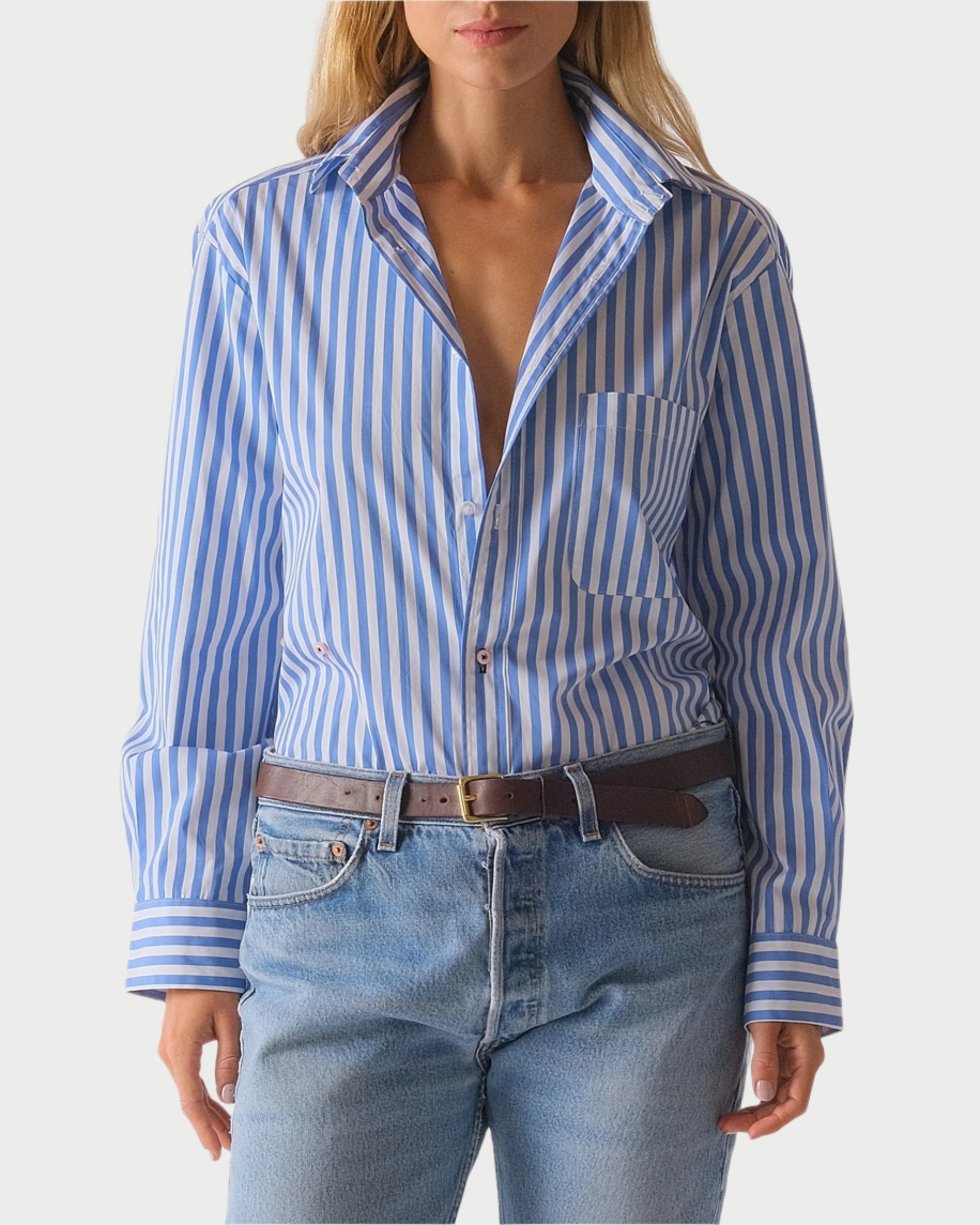 The person sports The Daily Shirt by WEAR CISSA, a blue and white striped Egyptian cotton piece, partially unbuttoned. It's paired with light blue jeans and accessorized with a visible brown leather belt. The look is completed against an unobtrusive background.