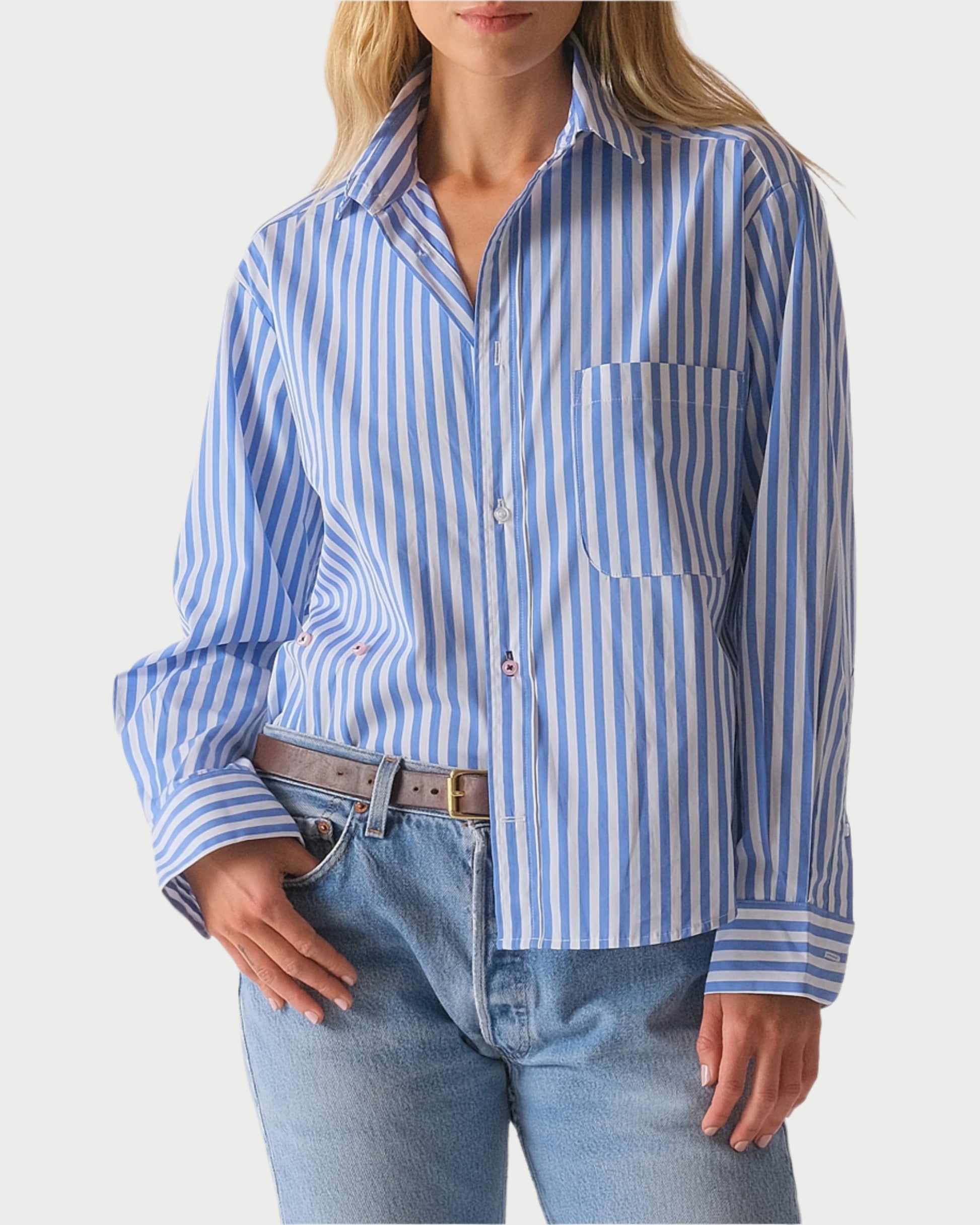 A person is styled in The Daily Shirt by WEAR CISSA, featuring blue and white vertical stripes, crafted from soft Egyptian cotton with long sleeves and a chest pocket. It's paired with light blue jeans and a brown belt, tucked into the jeans on one side for a versatile look.