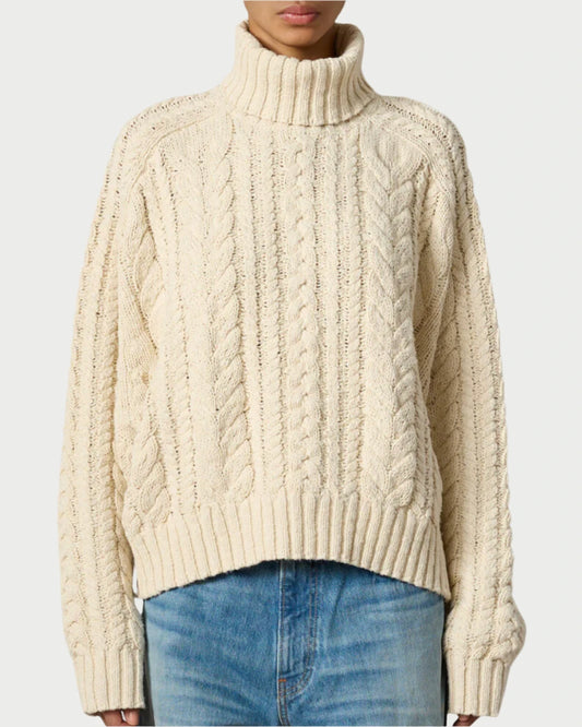 A person is wearing an oversized, cream-colored Cable Turtleneck Sweater by Denimist. The sweater features a high neck and long sleeves, made from cable-knit cotton. They have paired it with light blue denim jeans, and the background is plain white.