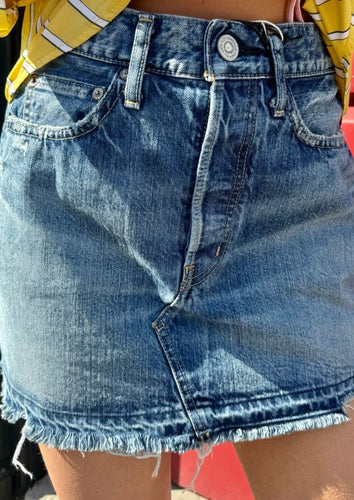Close-up of a person wearing a blue Moussy Mini Denim Skirt with a fringe hem, front pockets, and a button closure. The person is also wearing a yellow and white striped top, partially visible. Perfect for a casual lunch outfit or summer dinner date. The background is out of focus.