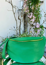 Load image into Gallery viewer, A bright green Crossbody Bag from DEBBIE KATZ with a gold zipper and a matching green strap sits on a glass table. The background features blooming pink flowers, green foliage, and bare branches, creating a contrasting natural scene behind the bag, perfect for traveling in style.