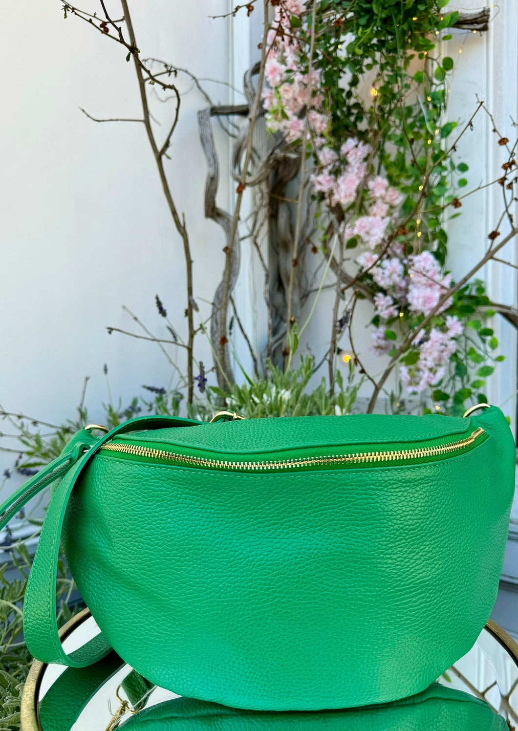 A bright green Crossbody Bag from DEBBIE KATZ with a gold zipper and a matching green strap sits on a glass table. The background features blooming pink flowers, green foliage, and bare branches, creating a contrasting natural scene behind the bag, perfect for traveling in style.