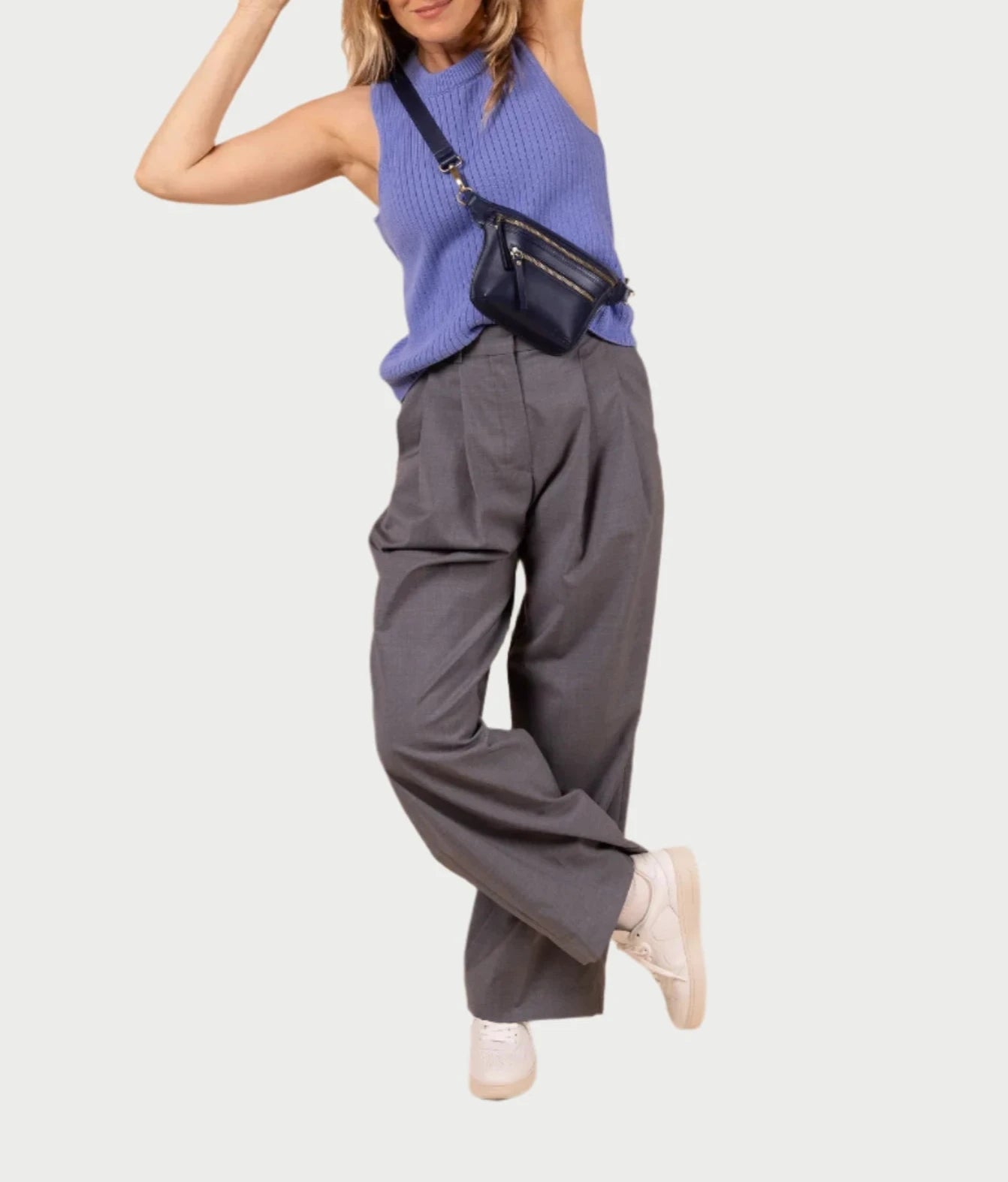 Dressed in a sleeveless blue top, gray wide-leg trousers, and white sneakers, a person poses with one leg raised. They sport O MY BAG's Beck's Bum Bag in Midnight Blue across the chest, with their arms raised against a plain background.