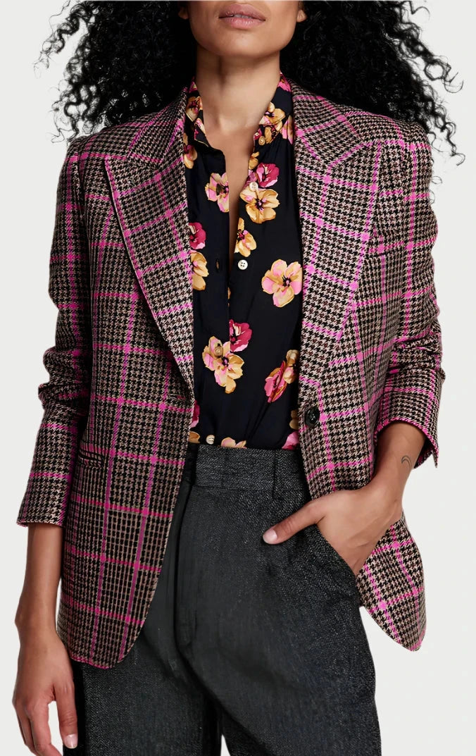 A person with long, curly hair stands confidently, wearing a Smythe Pink Glencheck 90's Blazer with rolled-up sleeves over a black floral shirt, and gray trousers. One hand is in their pocket, making this fall must-have outfit effortlessly stylish as they look directly at the camera.